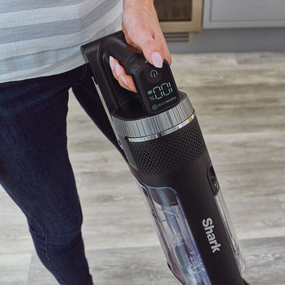 A person holding a Shark cordless stick vacuum cleaner in a home setting with a light wood floor.